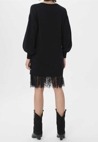 Svart stickad klänning med ballongärmar, prydd med en spetsdetalj vid nederkanten. Texturerad, midi-längd design som kombineras med svarta ankelstövlar.