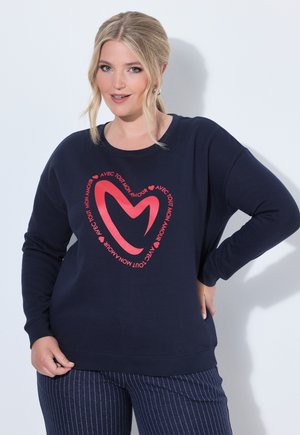 Lyshåret kvinde iført en marineblå sweatshirt med et rødt hjerte-design og fransk tekst, kombineret med marineblå bukser med striber, poserer med den ene hånd på hoften.