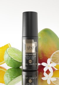 Flacon spray noir format voyage étiqueté « ghd curly ever after » avec des agrumes, de la mangue et des fleurs blanches autour.