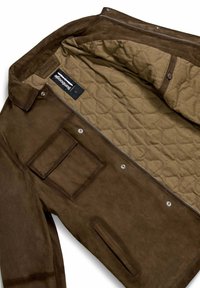Bruine suède jas met een gewatteerde beige voering, voorkant met rits, drukknoopjes en vierkante pocketdetails aan de buitenkant. Label zichtbaar aan de binnenkant.