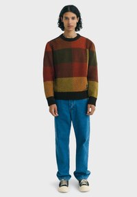Pull à col rond multicolore avec des carreaux rouges, verts et jaunes présentant des motifs diagonaux, associé à un jean bleu et des chaussures noires.