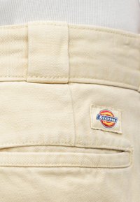 Beżowe spodnie z płótna z szerokim paskiem w pasie, tylną kieszenią i tkanym znakiem z czerwono-niebieskim logo „Dickies”.