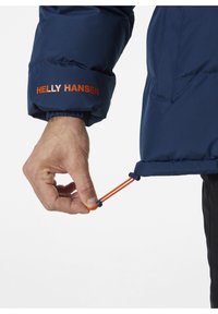 Chaqueta aislante de color azul marino con puños elásticos, que presenta un destacado logo de "Helly Hansen" en color naranja. Cuerda ajustable en el dobladillo.