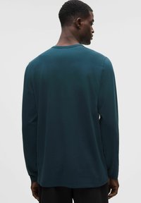 Dlhý rukáv košele v tmavej tyrkysovej farbe, vyrobená z mäkkej bavlny, s okrúhlym výstrihom a voľným strihom, s hladkou textúrou a postrannými rozparkami.