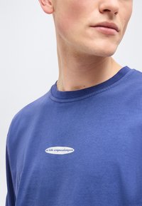 Κοντινό πλάνο ενός ατόμου που φοράει μπλε μπλουζάκι με στρογγυλή λαιμόκοψη και μικρό λευκό οβάλ λογότυπο που γράφει "arkh copenhagen" στο στήθος.