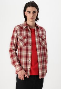 Rot- und cremefarbener karierter Flanellhemd mit zwei Brusttaschen, schwarzen Knöpfen, über einem roten T-Shirt getragen, vor einem weißen Hintergrund präsentiert.