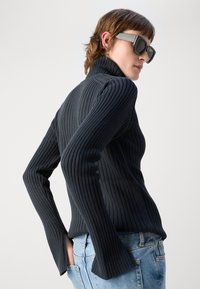 Maglione a collo alto nero a coste con maniche lunghe, caratterizzato da una silhouette aderente e dettagli con polsini svasati, abbinato a jeans azzurri chiari.