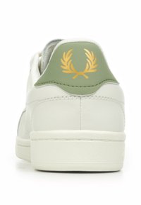 Sneaker bianca con tallone verde e un emblema di alloro dorato. Materiale in pelle liscia e suola testurizzata, design minimalista.