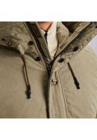 Béžová vypchávkovaná bunda s kapucňou, s predným zipsom, čiernymi gombíkmi a hnedými šnúrkami. Textúrová tkanina s viditeľným šitím.