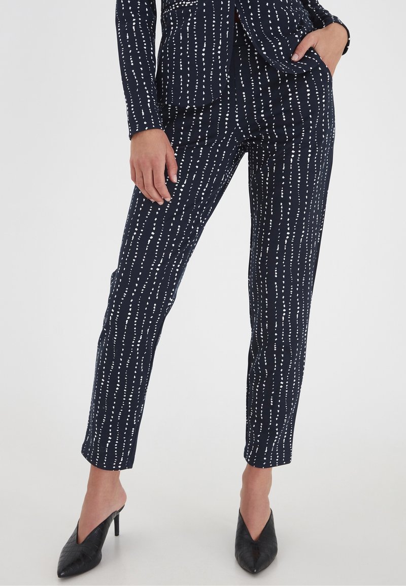 Pantalones navy con rayas verticales blancas, diseñados en un corte ajustado con bolsillos, combinados con zuecos de tacón negro. Textura de tela suave.