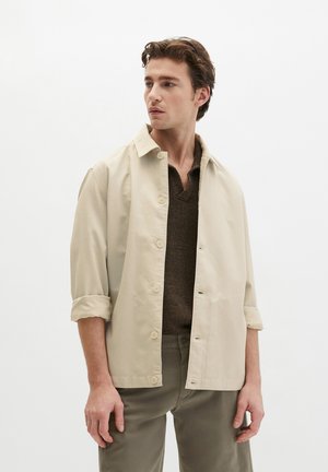 Junger Mann trägt eine beige Jacke mit Knöpfen über einem braunen Strickpullover und olivgrünen Hosen, blickt nach links vor einem einfarbigen weißen Hintergrund.