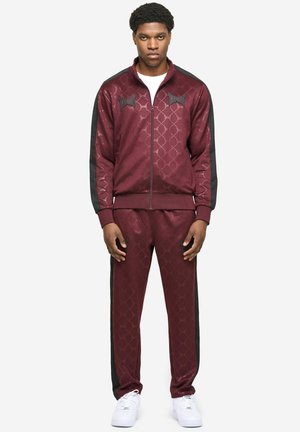 Maroon värvi trennikomplekt kuuekandilise mustriga ning mustade küljeribadega. Lukuga jope ja sobivad püksid, valmistatud siledast kangast. Valged jalanõud.