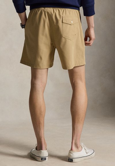 Muž v béžových šortkách s zadným vreckom, tmavomodrej dlhorukávovej košeli a bielych slip-on topánkach stojí na šedom povrchu.