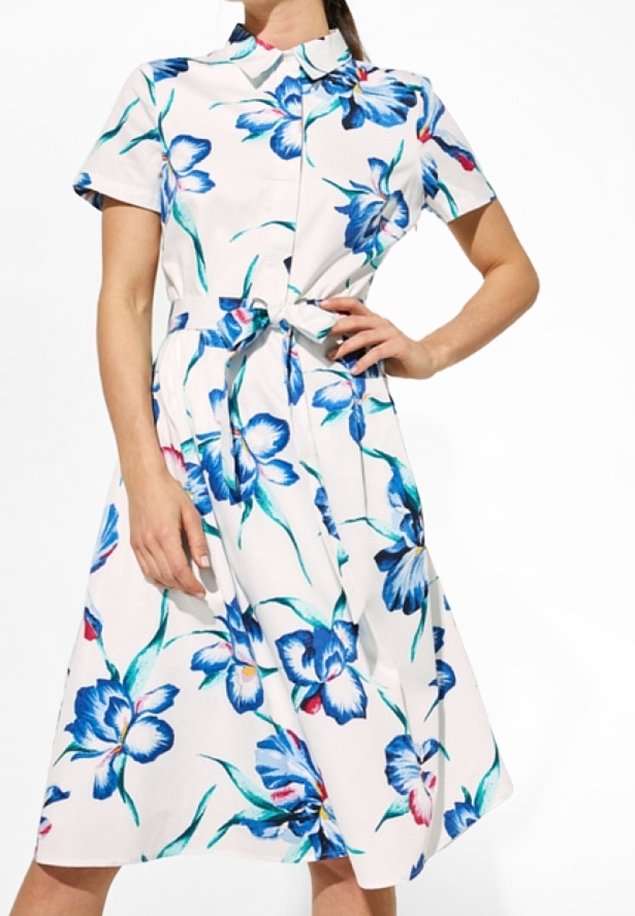 Femme portant une robe blanche à manches courtes avec des motifs floraux bleus et verts, cintrée à la taille avec une ceinture assortie en tissu.