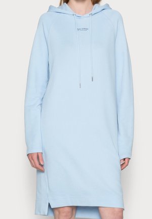 Žena v světle modrých dlouhých rukávech mikinových šatech s kapucí, šňůrkami a malým logem na hrudi, stojící na neutrálním pozadí.