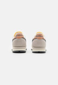 Zapatillas Reebok con parte superior blanca y gris, acentos de gamuza beige y un forro amarillo. Textura suave con una suela acolchada.