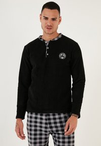 Camisa de manga larga de forro polar negro con un cuello a cuadros gris y botones, que presenta un parche redondo de logo en el lado izquierdo del pecho. Combinada con pantalones de cuadros.