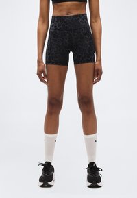 Γυναίκα που φοράει αθλητικά σορτς με μαύρο animal print και ψηλή μέση, μαύρο αθλητικό σουτιέν, λευκές ριμπ κάλτσες και μαύρα παπούτσια τρεξίματος σε λευκό φόντο.