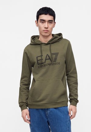 Ung mand iført en olivengrøn EA7 Emporio Armani hættetrøje og blå jeans, stående mod en ensfarvet lys baggrund.