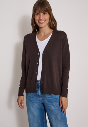 Femme souriante portant un cardigan marron boutonné par-dessus une chemise blanche et un jean bleu, debout devant un fond clair uni.