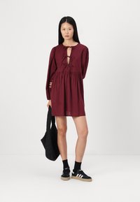 Vestido bordô com mangas longas, frente em formato de olho de fechadura e cintura pregueada. Combinado com ténis pretos e uma mala de mão preta texturizada.