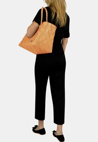 Orange Woven Tote Bag mit strukturiertem Oberflächen, schlanken Trägen und silbernem Logo-Akzent, kombiniert mit einem schwarzen Jumpsuit und schwarzen Slippern.