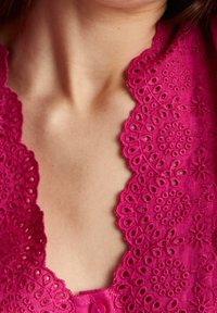Blusa di cotone rosa ricamata con scollatura a fodera e motivi floreali, dotata di chiusura frontale a bottoni. Visibile un dettaglio di tessuto testurizzato.