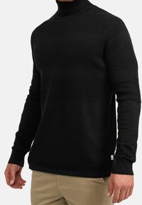 Svart polotröja med texturerade horisontella ränder, ribbade ärmslut och krage, samt en slät, figursydd design.