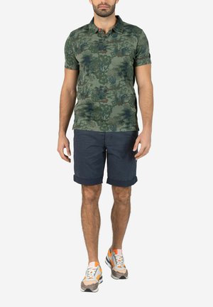 Polohemd aus grünem Camouflage-Stoff, mit einem Kragen, kurzen Ärmeln, kombiniert mit navyblauen Shorts und grauen/orangen Turnschuhen.
