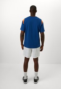 Kortermet blå T-skjorte med oransje detaljer, kombinert med hvite shorts med et subtilt mønster. Svarte sneakers og hvite sokker.