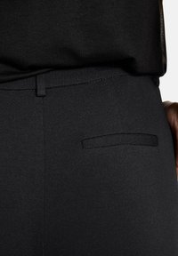 Close-up van een persoon in zwarte, op maat gemaakte broeken met een achterzak en een bijpassend zwart shirt dat erin gestopt is.