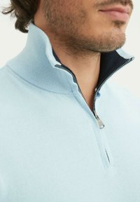 Pull-over bleu clair avec col zippé à quart, doté d'un col côtelé et d'une doublure intérieure contrastante en marine. Présente une fermeture éclair argentée et une texture douce.