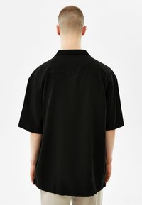 Persona con capelli corti che indossa una maglietta nera larga a maniche corte e pantaloni chiari, in piedi girata di spalle su uno sfondo bianco.