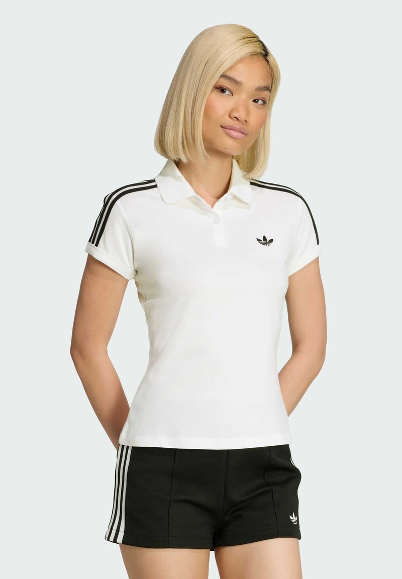Camisa polo blanca con rayas negras en las mangas y un pequeño logo negro. Combinada con pantalones cortos negros que tienen tres rayas blancas.