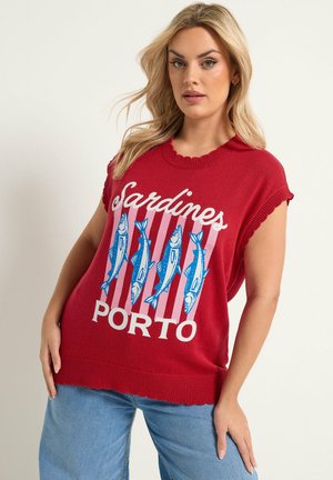 Mujer rubia con un suéter sin mangas rojo con gráficos azules de sardinas y el texto "Sardines PORTO", combinado con vaqueros azul claro.