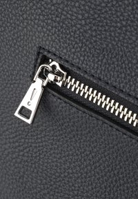Materiale nero testurizzato con una cerniera e un tirante in argento. Presenta una chiusura rettangolare con cerniera e dettagli di cucitura prominenti.