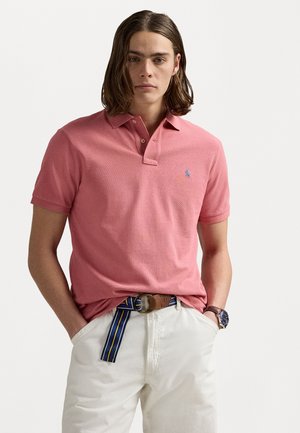 Polo de color rosa claro, de mangas cortas, con cuello y un pequeño logo azul en el lado izquierdo del pecho. Combinado con pantalones blancos y un cinturón a rayas.