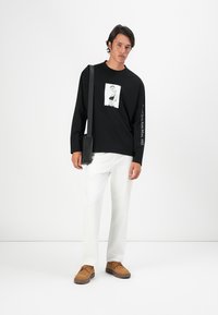 Zwarte lange mouwen shirt met een grafische print, witte tekst op de mouw, gecombineerd met witte broek en bruine schoenen, met een kleine zwarte tas.