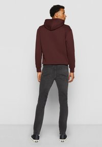 Bordeauxrote Kapuzenjacke, graue Slim-Fit-Jeans, lässige Sneaker. Schlichte Gestaltung ohne sichtbare Muster oder Hardware. Weiches Material, lockere Passform.