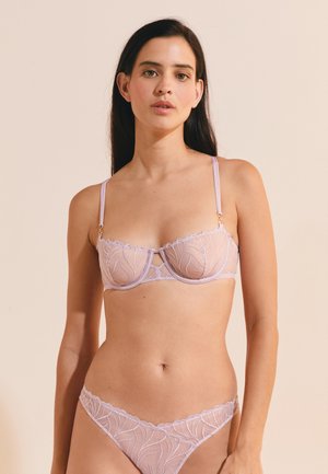 Donna che indossa un reggiseno in pizzo trasparente color lavanda chiaro e mutandine abbinate su uno sfondo beige uniforme.