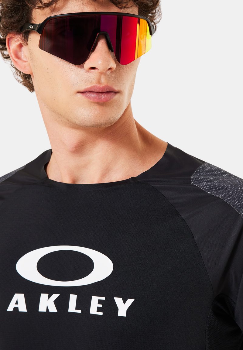 Homme portant un t-shirt noir Oakley et de grandes lunettes de sport réfléchissantes rouge-orange, regardant vers la droite, sur un fond clair uni.