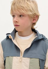 Veste zippée présentant une palette de couleurs bleu clair et crème avec des accents verts, fabriquée en tissu texturé et léger.