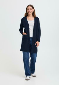 Cardigan long marine à ouverture sur le devant, avec deux poches plaquées et des poignets côtelés, associé à un haut gris clair et un jean bleu.