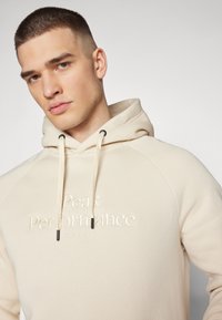 Beige kapucnis pulóver, texturált anyaggal, bordázott mandzsettákkal, feketére színezett húzózsinórokkal és hímzett logóval az elején.