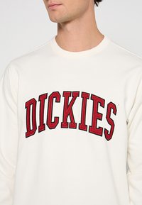 Felpa bianca con logo "DICKIES" rosso ricamato ad arco, con scollo rotondo e polsini a costine.
