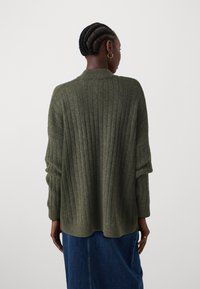 Grønn ribbestrikket genser med høy hals og oversized ermer, laget av et mykt strikkemateriale, kombinert med et jeanskjørt.