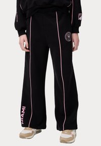 Pantalones de chándal negros con franjas laterales rosas, cinturilla elástica y un logo de Miami. Fabricados en tela suave. Presentan un diseño de pierna ancha.