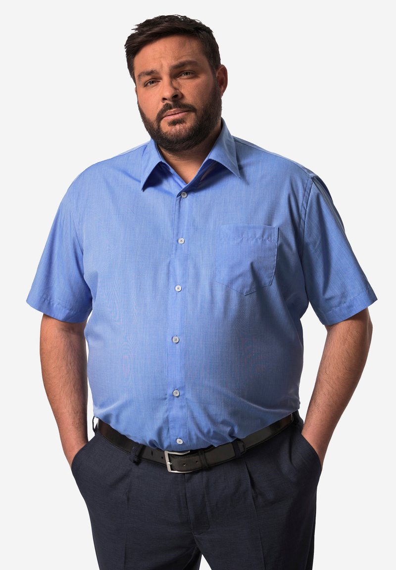 Hombre con el pelo corto y oscuro y barba, lleva una camisa azul de manga corta con botones y pantalones oscuros, manos en los bolsillos, mirando al frente.