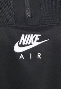 Svart tyg med vit broderad Nike Air-logotyp under en svart dragkedja och meshpaneler.