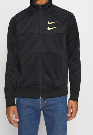Mann trägt schwarze Sportjacke mit Reißverschluss und zwei gelben Nike-Swoosh-Logos auf der Brust sowie blaue Jeans.